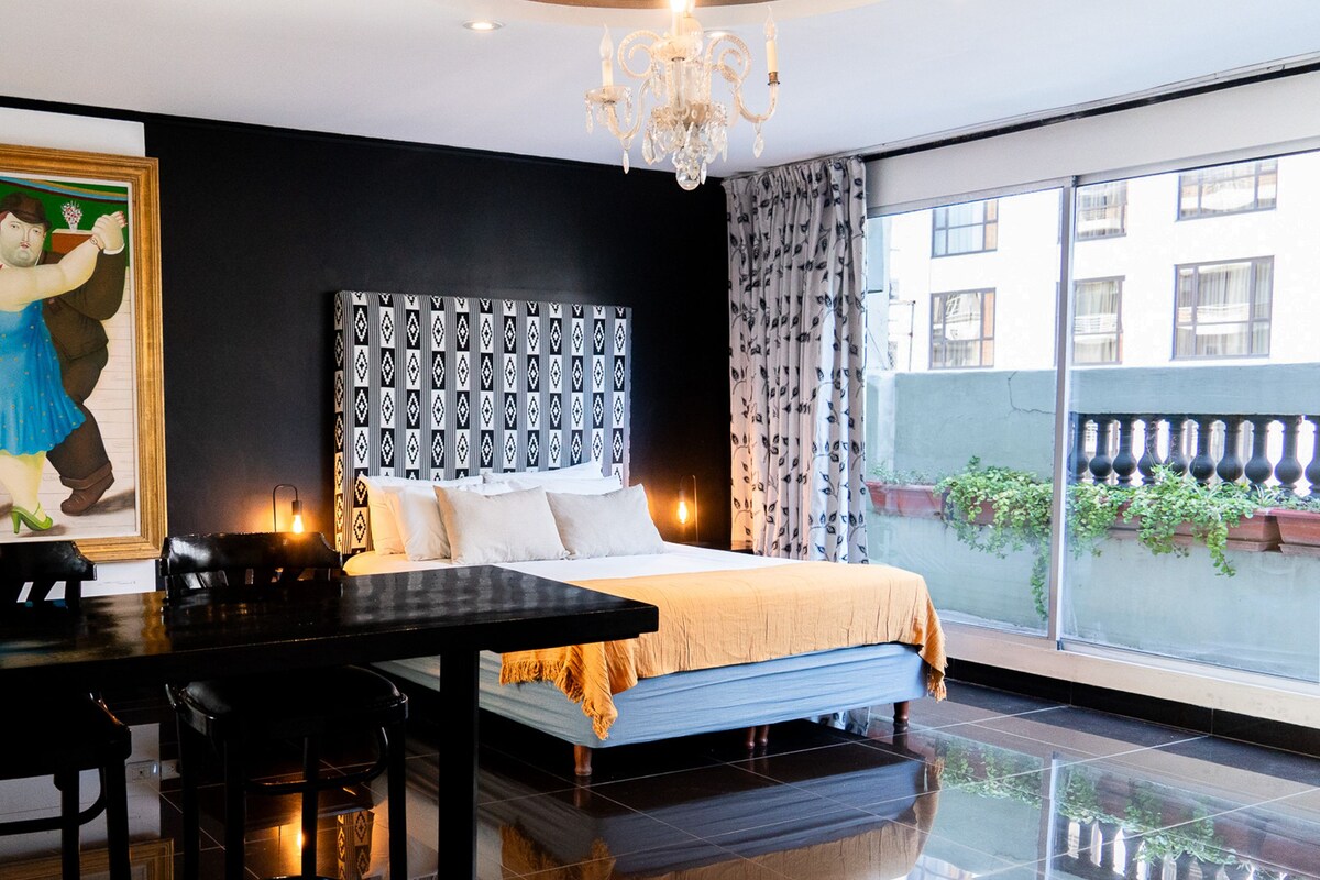 Quarto de hotel com cama grande, mesa de jantar, lustre e vista para o exterior.