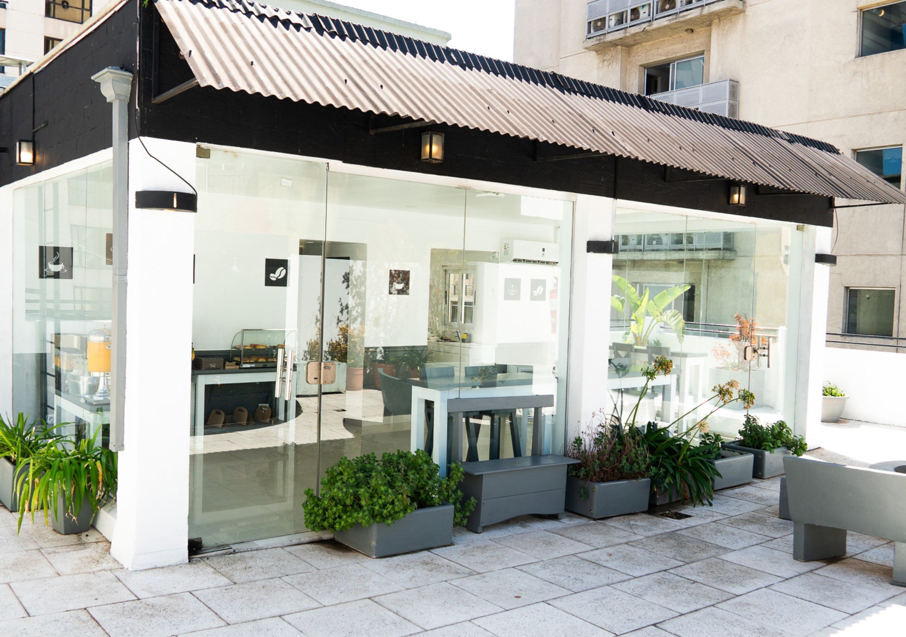 A modern outdoor cafe with glass walls, seating, and potted plants.