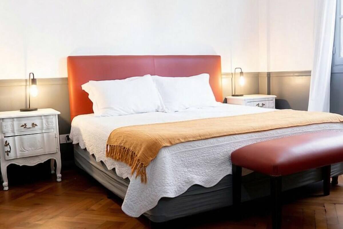 Un dormitorio ordenado con cama, mesitas de noche, un banco y una lámpara.