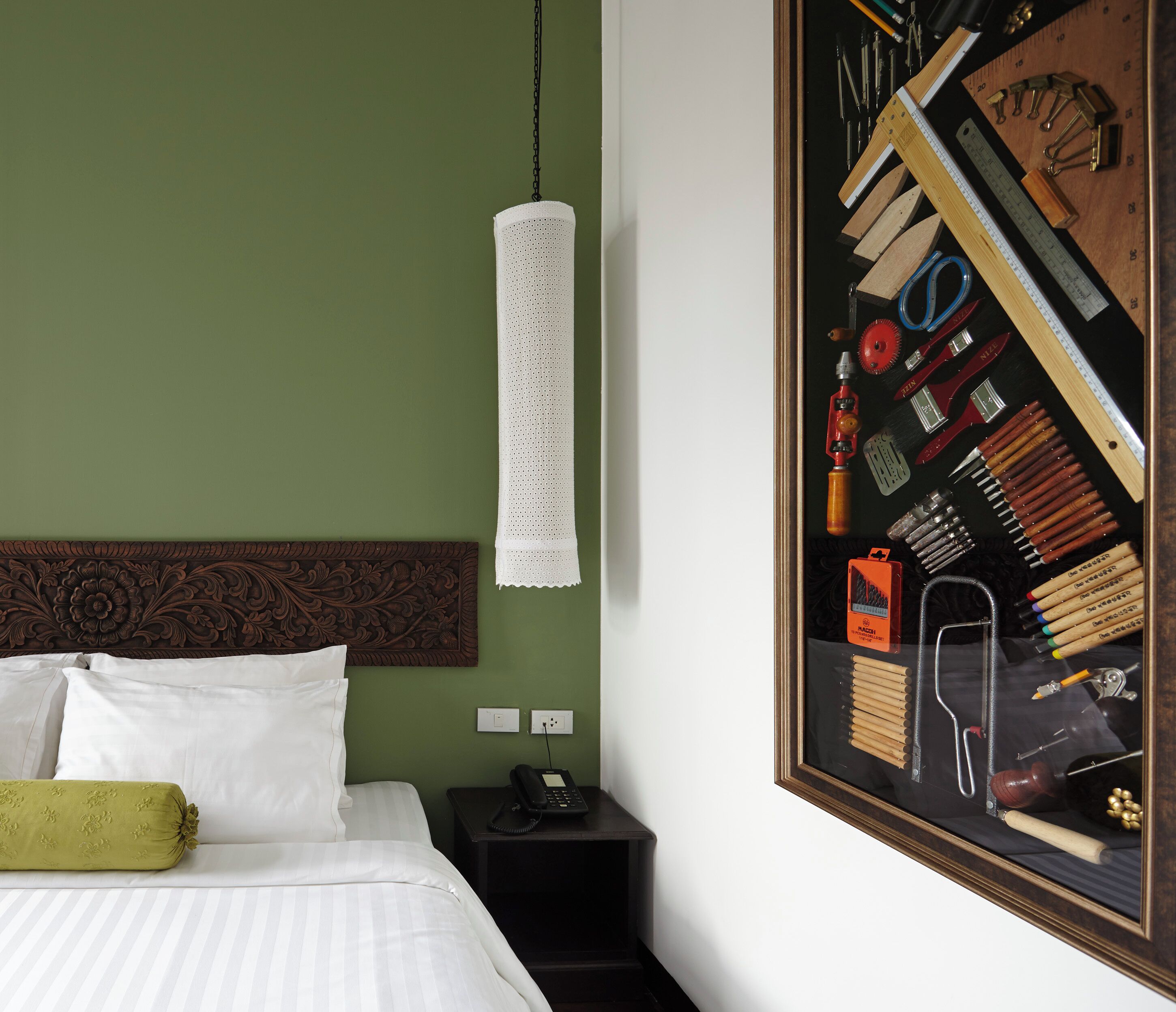 A neatly made bed with white linens and a green pillow, a wooden headboard, a hanging white towel, a nightstand with a phone, and a framed picture of tools on the wall.
