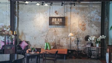 A rustic interior with a sign reading "On On hotel," a bench, chairs, a table with a vase of flowers, and a fan against a textured wall.