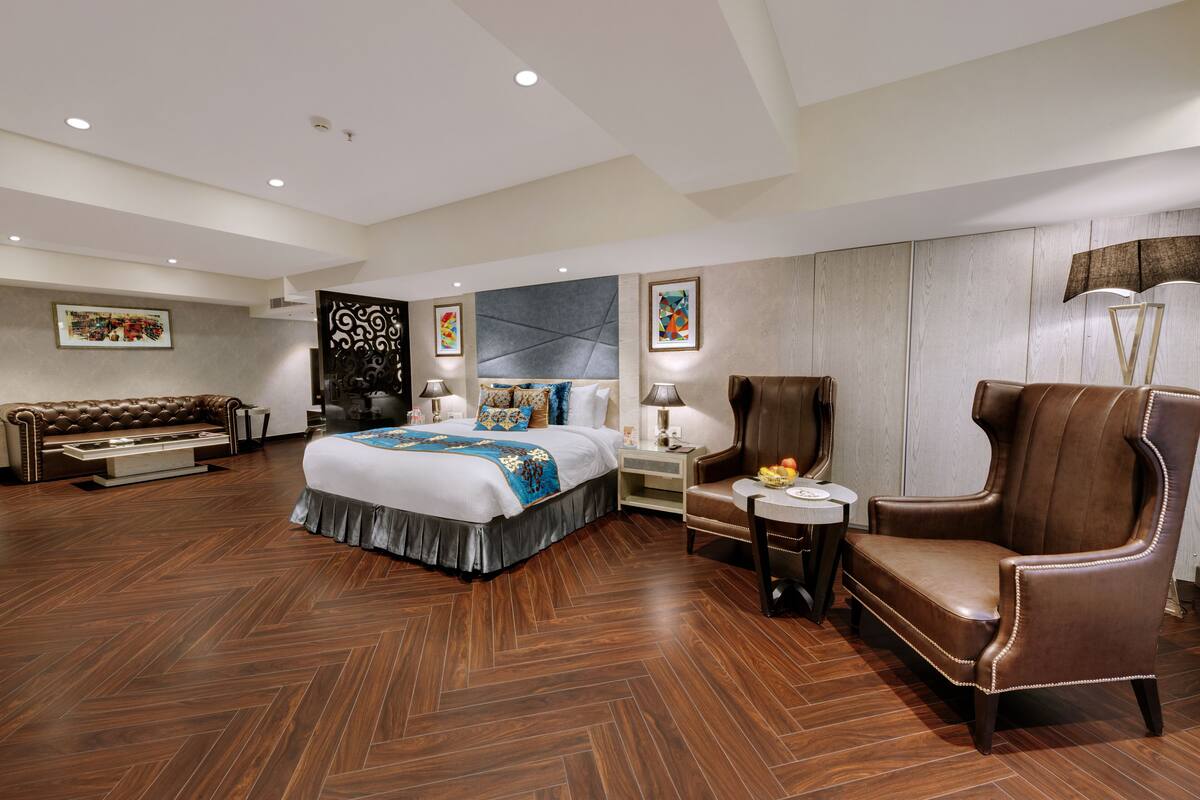 Amplia habitación de hotel con una cama grande, un sillón de cuero, un sofá y una pequeña mesa con una lámpara.