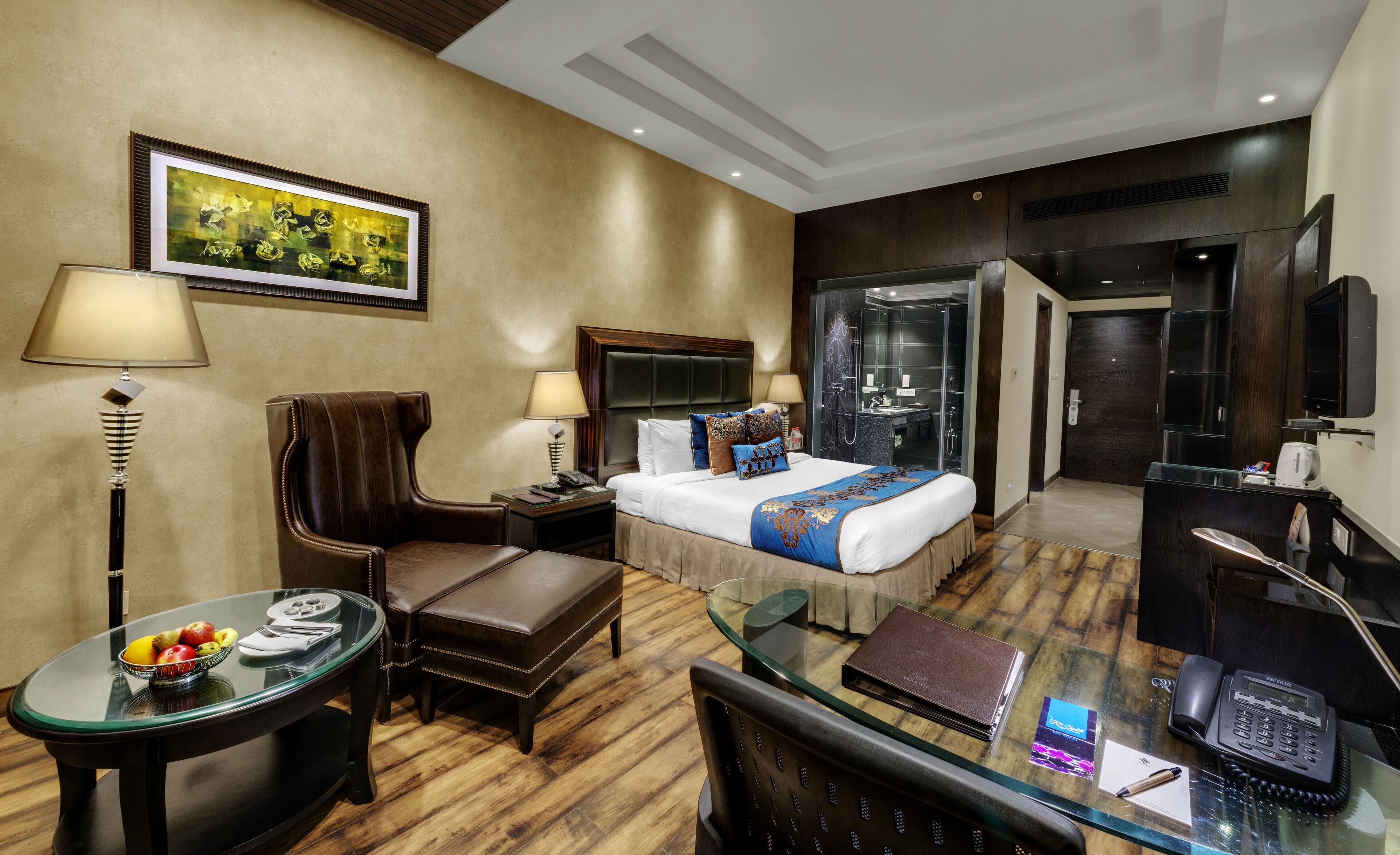 A hotel room with a bed, a desk with a telephone, a chair, a glass table with fruit, and a painting on the wall.
