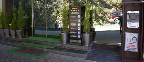 Entrada de hotel con un cartel que enumera precauciones por COVID-19, dispensador de desinfectante para manos y plantas en macetas.