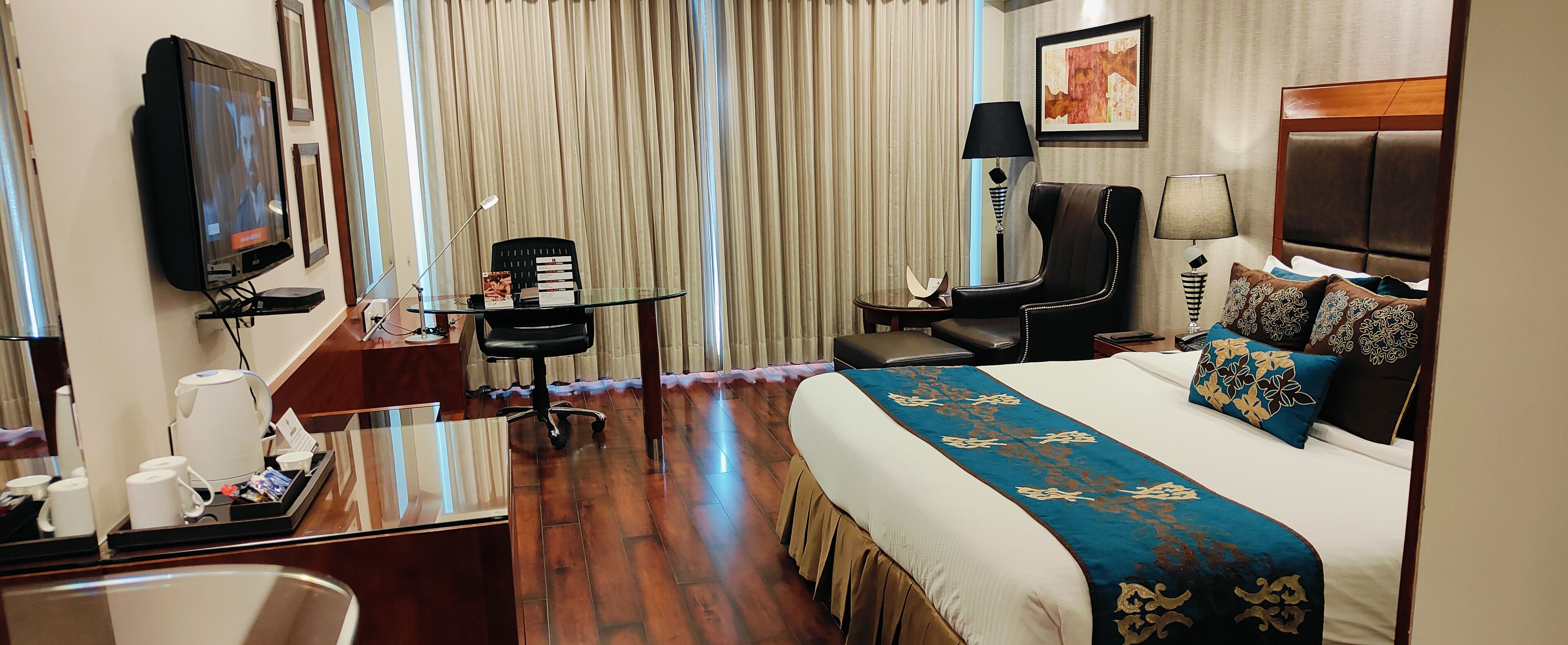 A hotel room with a bed, a desk with a computer, a chair, a TV, and a window with curtains.