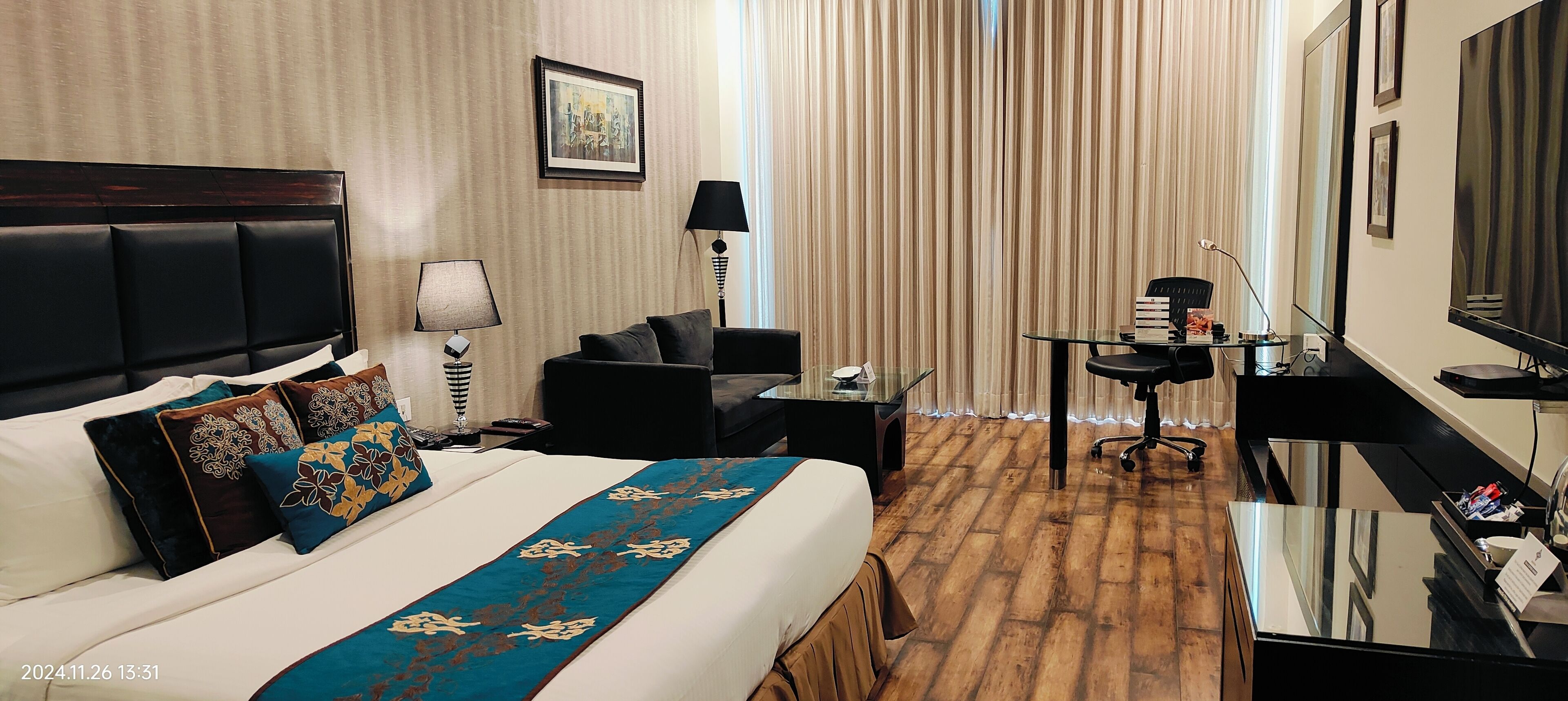 A hotel room with a large bed, a desk, a chair, a lamp, and a TV.
