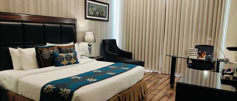 A hotel room with a large bed, a desk with a lamp, a chair, and a minibar.