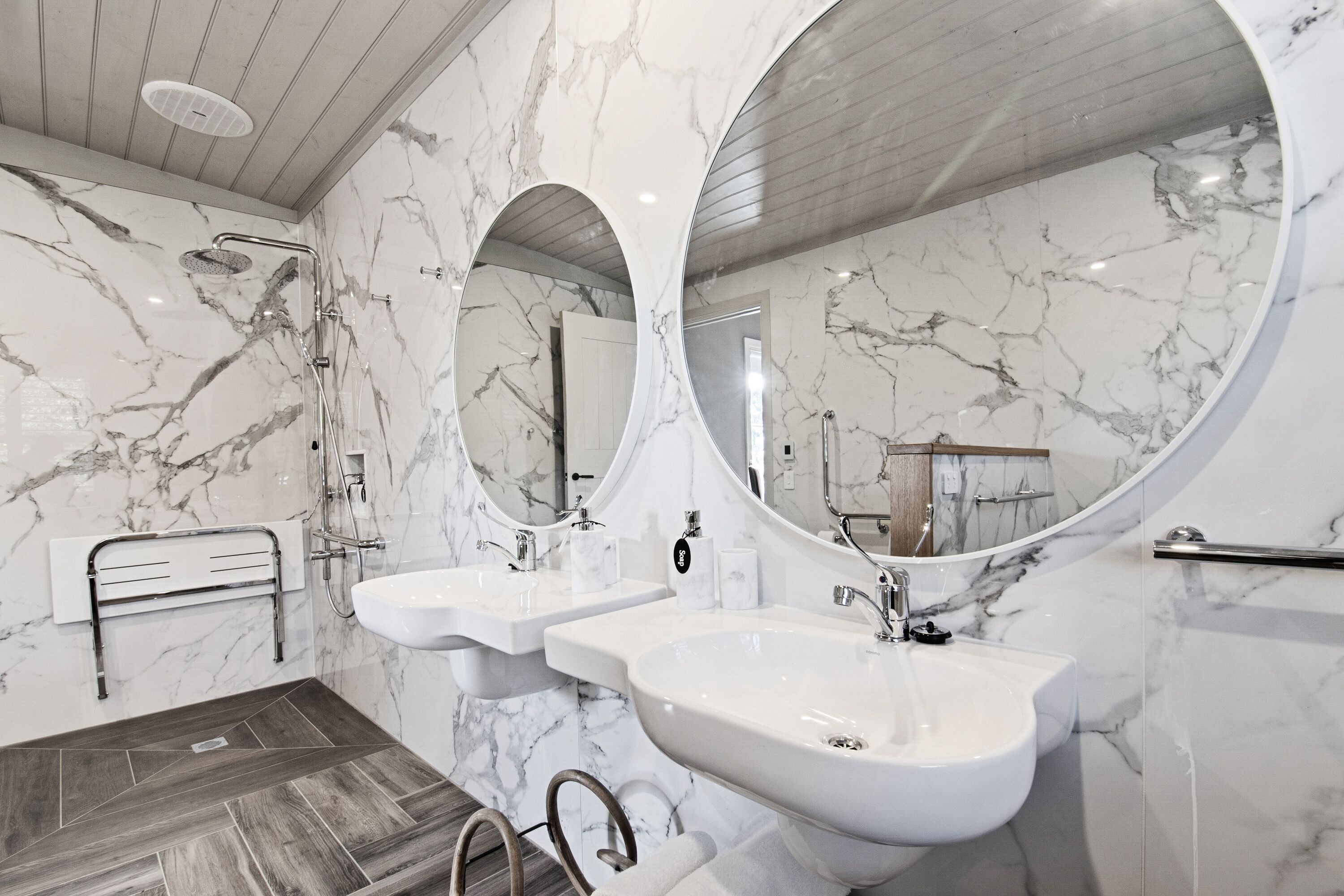 A modern bathroom with marble walls, two sinks, a shower, and a wooden floor.