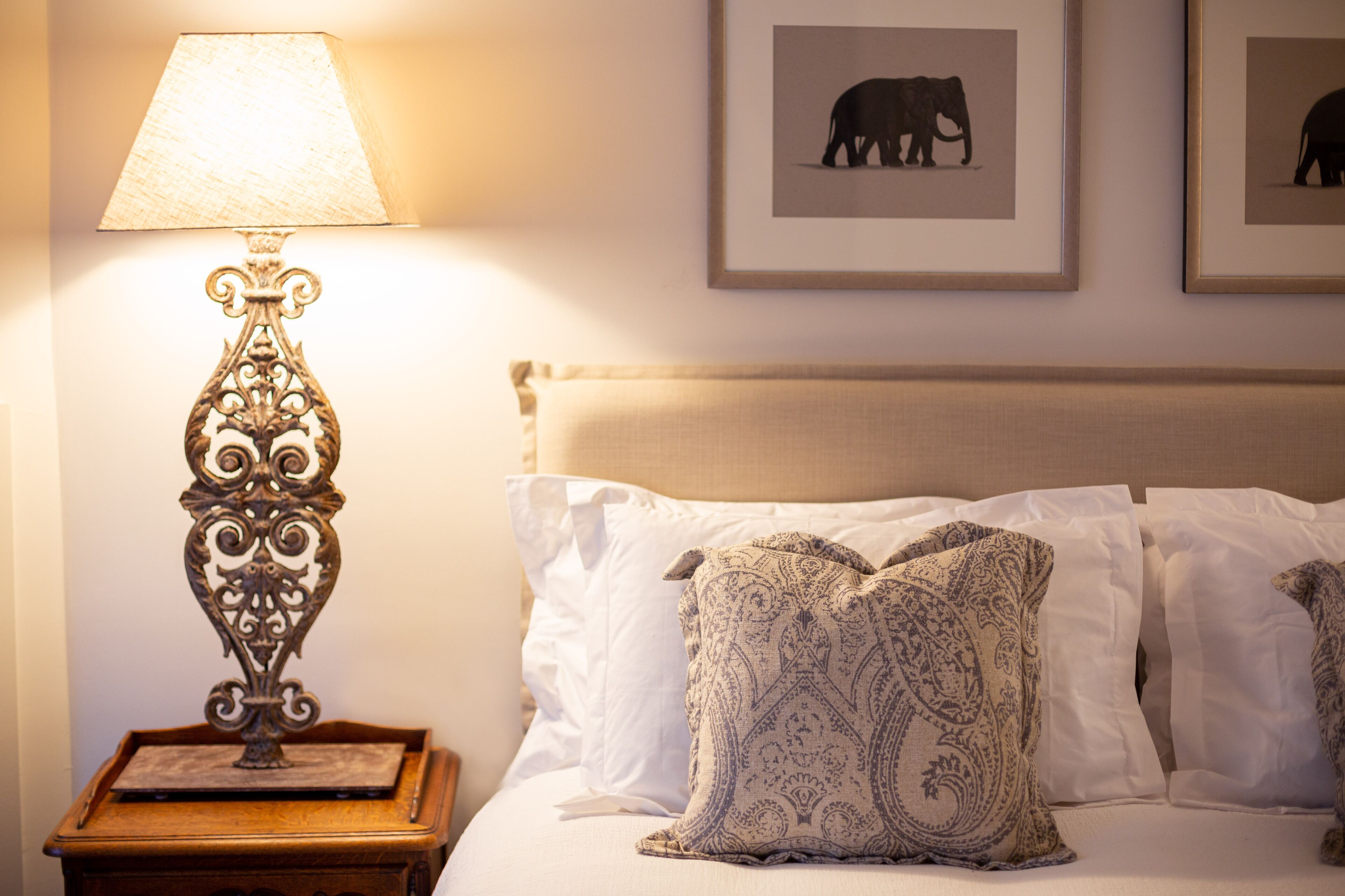 A neatly made bed with patterned pillows, a decorative table lamp, and framed elephant artwork on the wall.