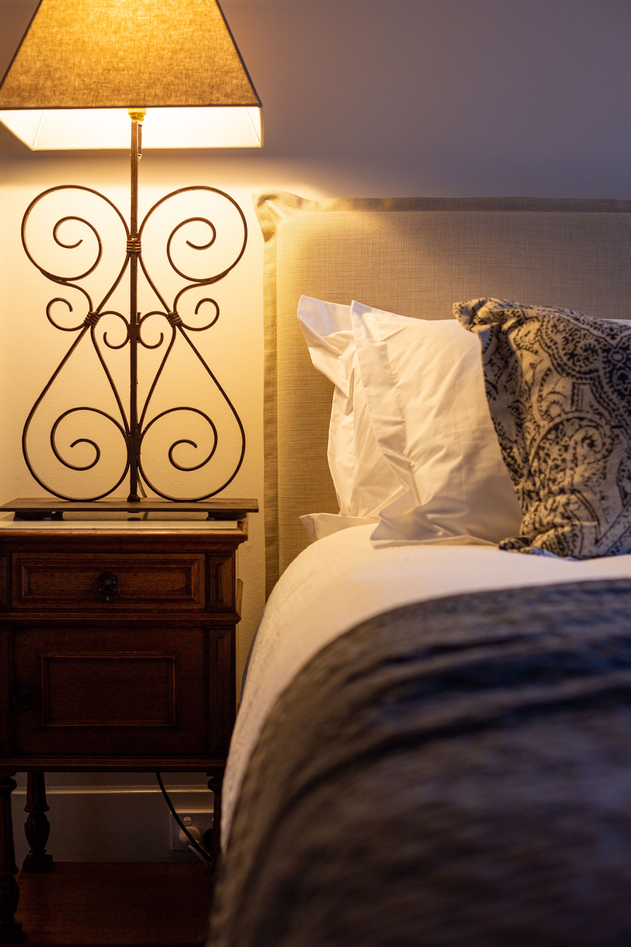 Een net opgemaakt bed met witte en gemêleerde kussens, een nachtkastje met een lamp en een houten bijzettafeltje.