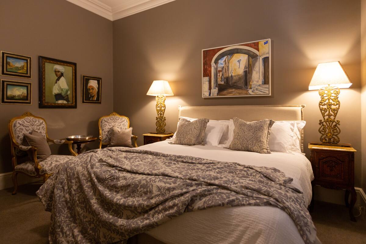 Habitación de hotel elegante con una cama grande, dos mesitas de noche, una silla y un cuadro en la pared.