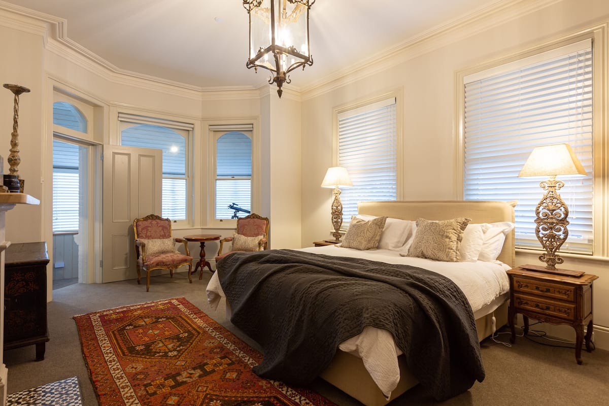 Un dormitorio con una cama grande, dos mesitas de noche, una silla, una alfombra y un candelabro.