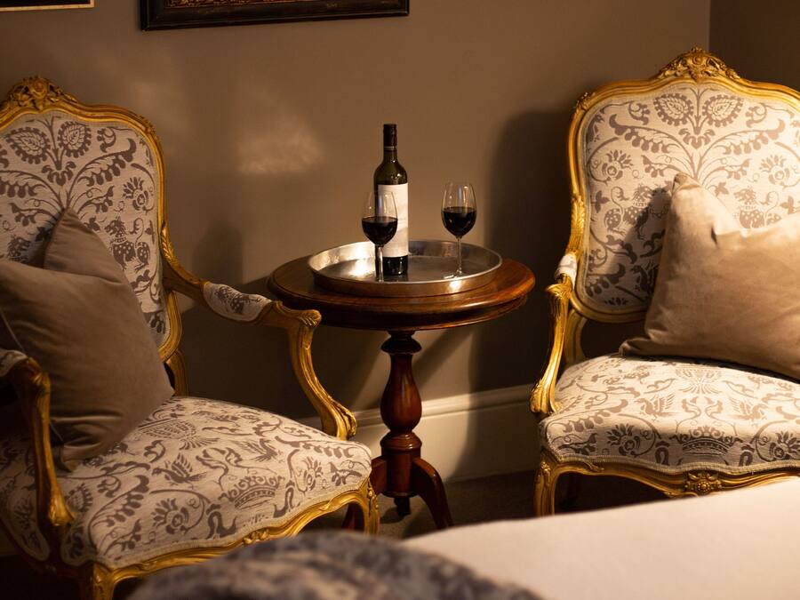 A room with two ornate chairs, a small round table with a bottle and glass of wine, and a bed with a patterned blanket.