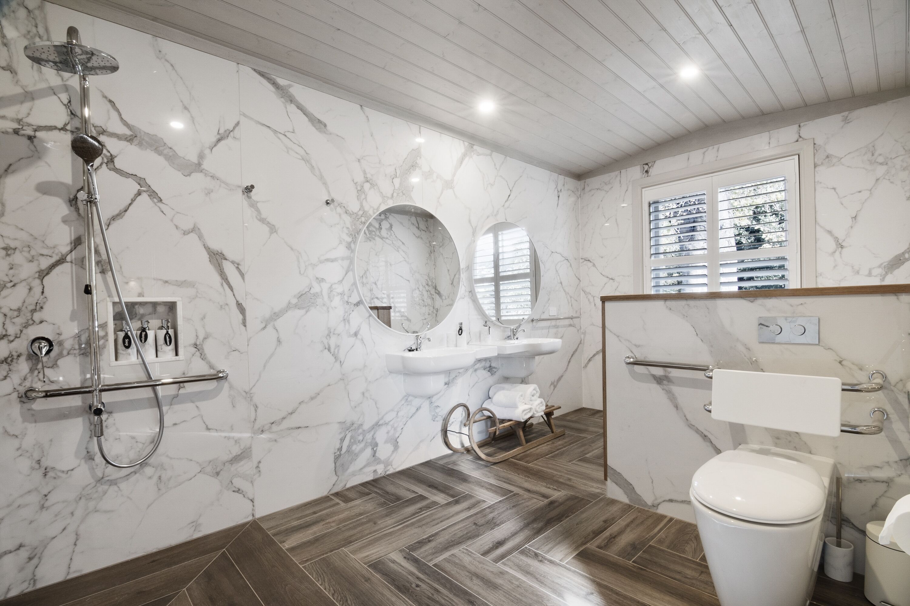 Un baño con pared de mármol, suelo de madera, ducha, inodoro y lavamanos.