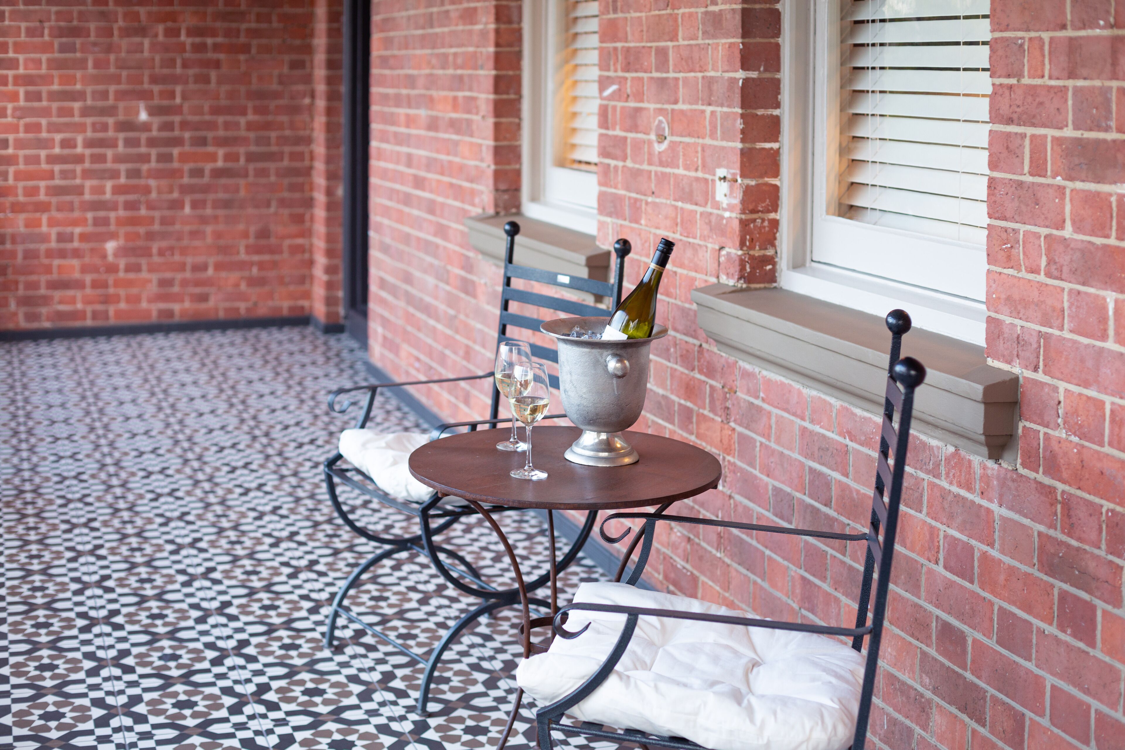 Un patio con una mesa y sillas de metal, una botella de vino y copas sobre la mesa.