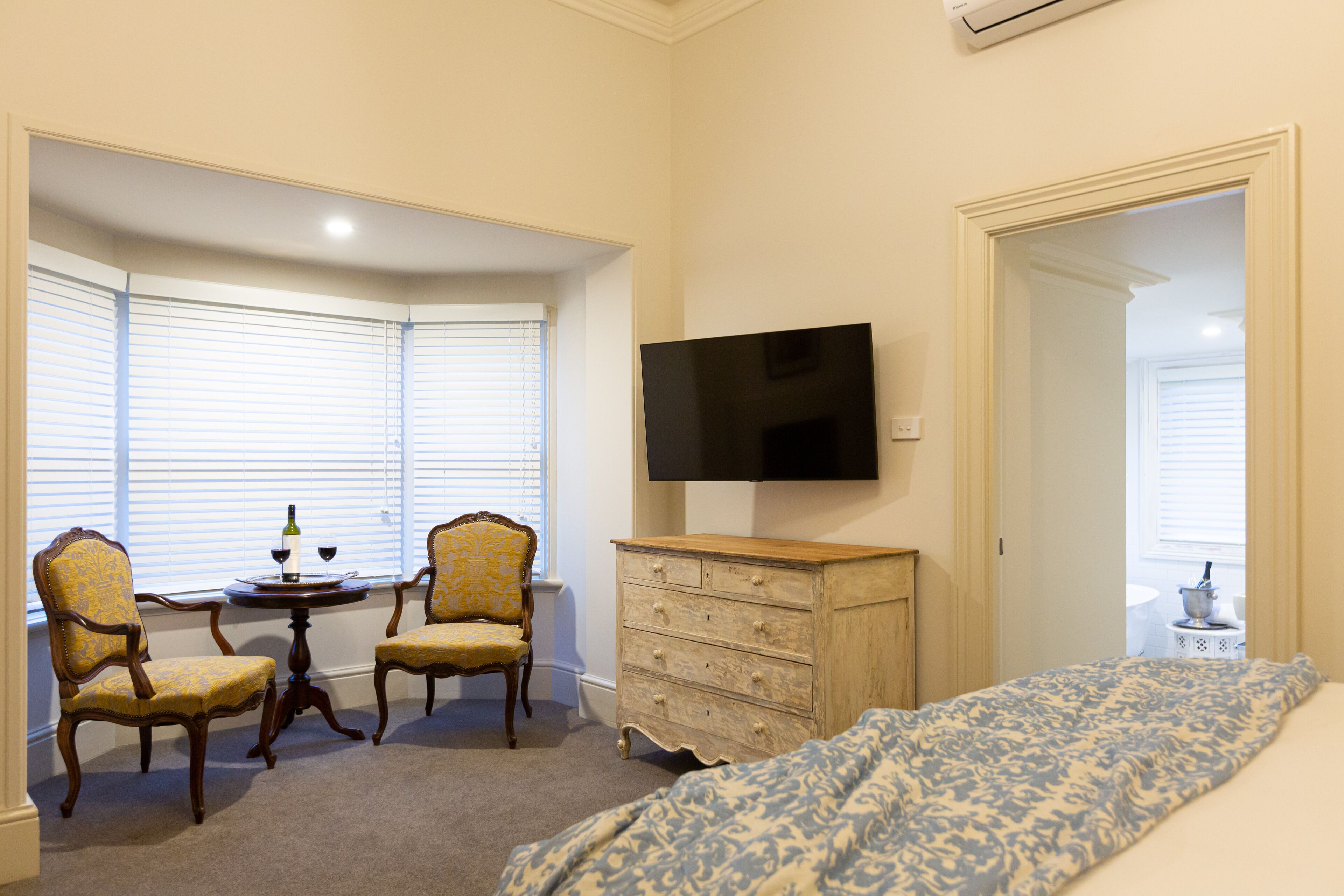 A bedroom with a bed, a dresser, a television, two chairs, a small table with a bottle, and a window with blinds.