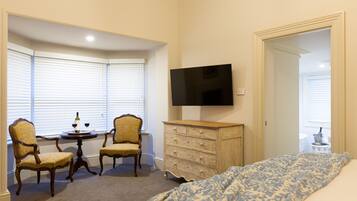 A bedroom with a bed, a dresser, a television, two chairs, a small table with a bottle, and a window with blinds.