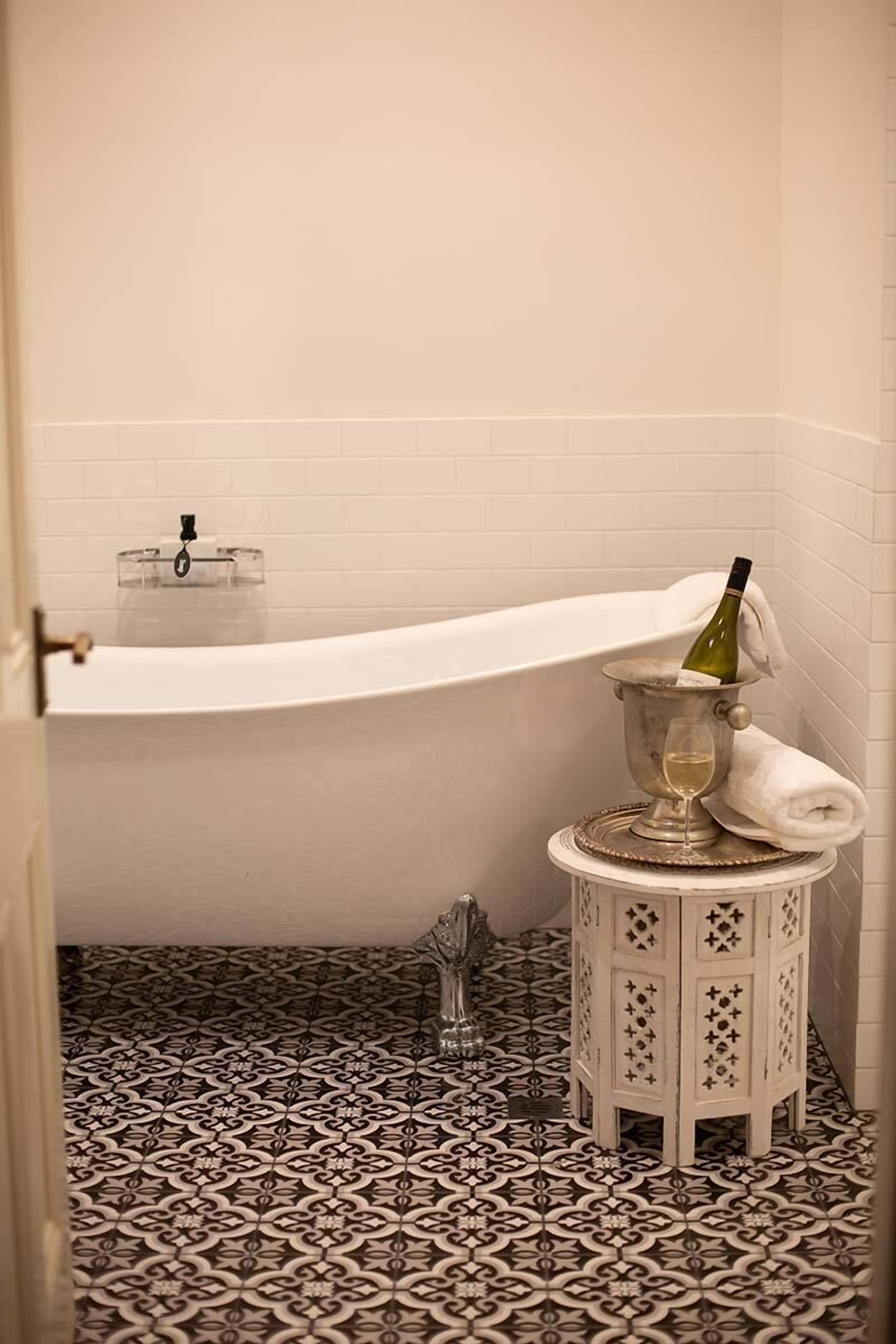 A bathroom with a white bathtub, a small round table with a wine bottle and glass, and patterned floor tiles.