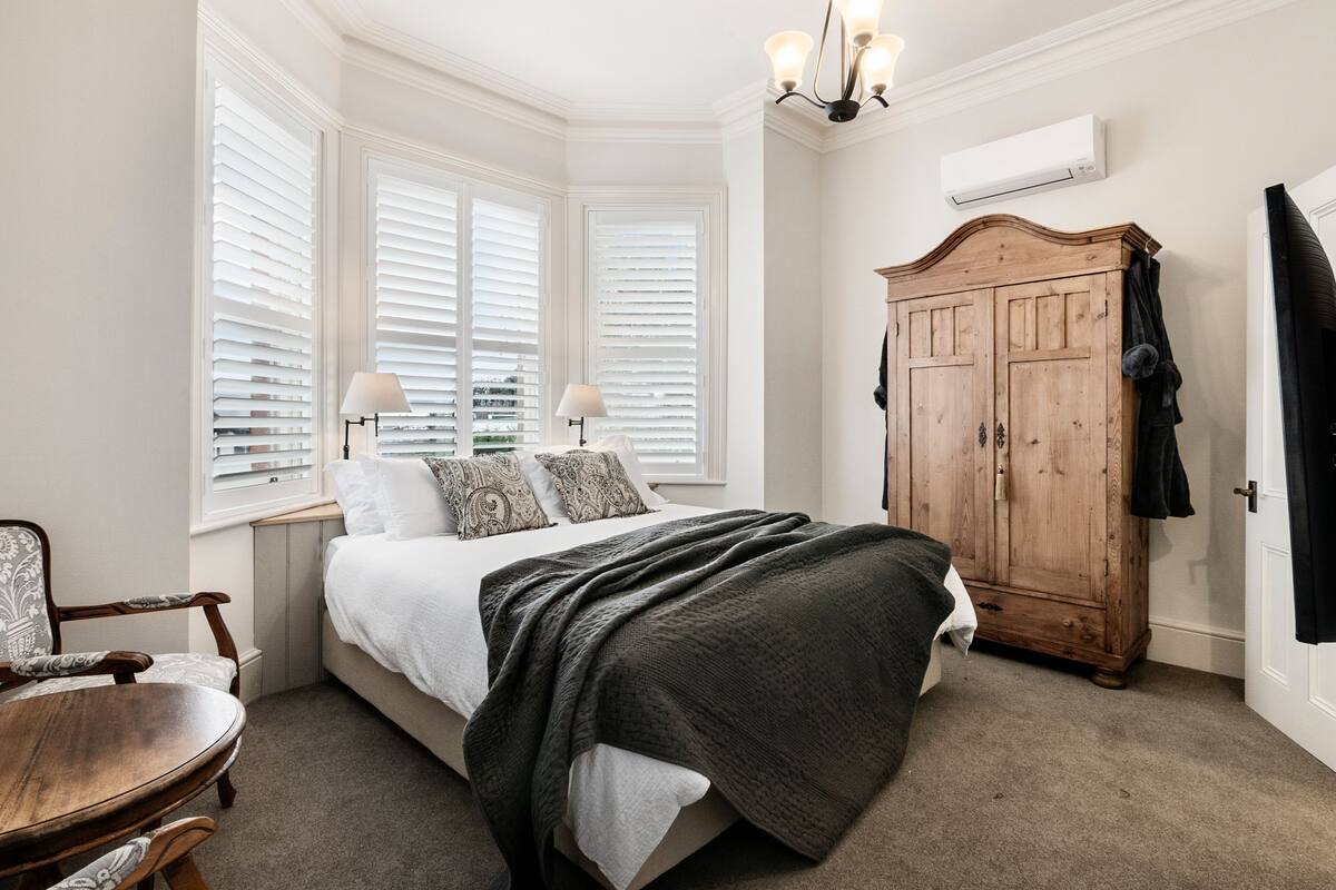 Un dormitorio con cama, armario de madera, silla, mesa y ventana con persianas.