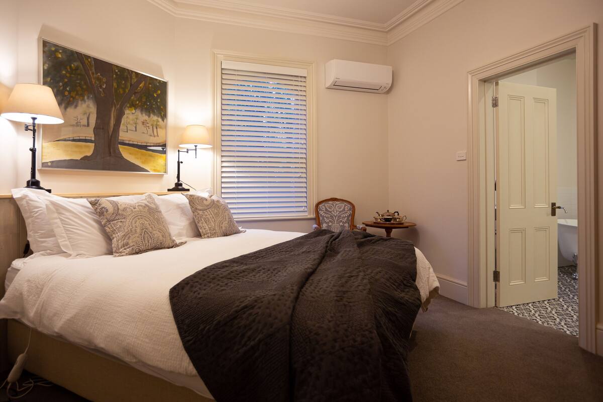 Un dormitorio con cama, dos lámparas de noche, un cuadro, una ventana con persianas, un equipo de aire acondicionado, una silla, una mesita con lámpara y una puerta que da al baño.