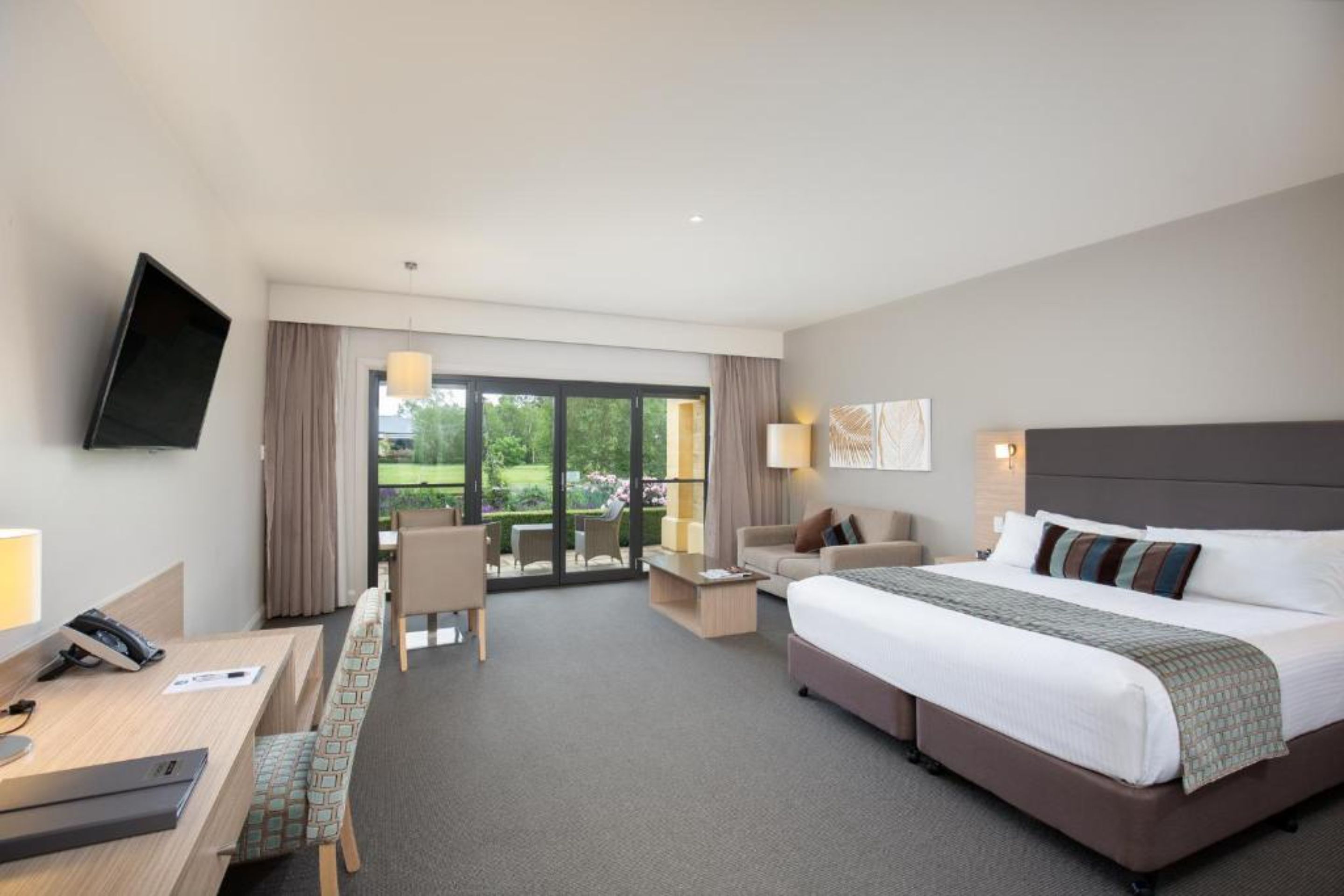 A hotel room with a large bed, a desk, a chair, a TV mounted on the wall, and a view of the outdoors through a sliding glass door.
