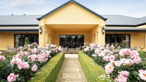 A building with a yellow facade, a central entrance, and a pathway flanked by pink flowers. - The Barn Accommodation (Ob Flat)