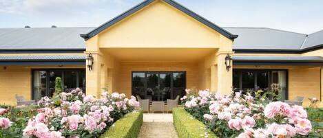 A building with a yellow facade, a central entrance, and a pathway flanked by pink flowers.