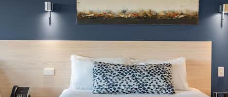 A hotel room with a wooden headboard, a bed with white and patterned pillows, a painting on the wall, and a bedside phone.