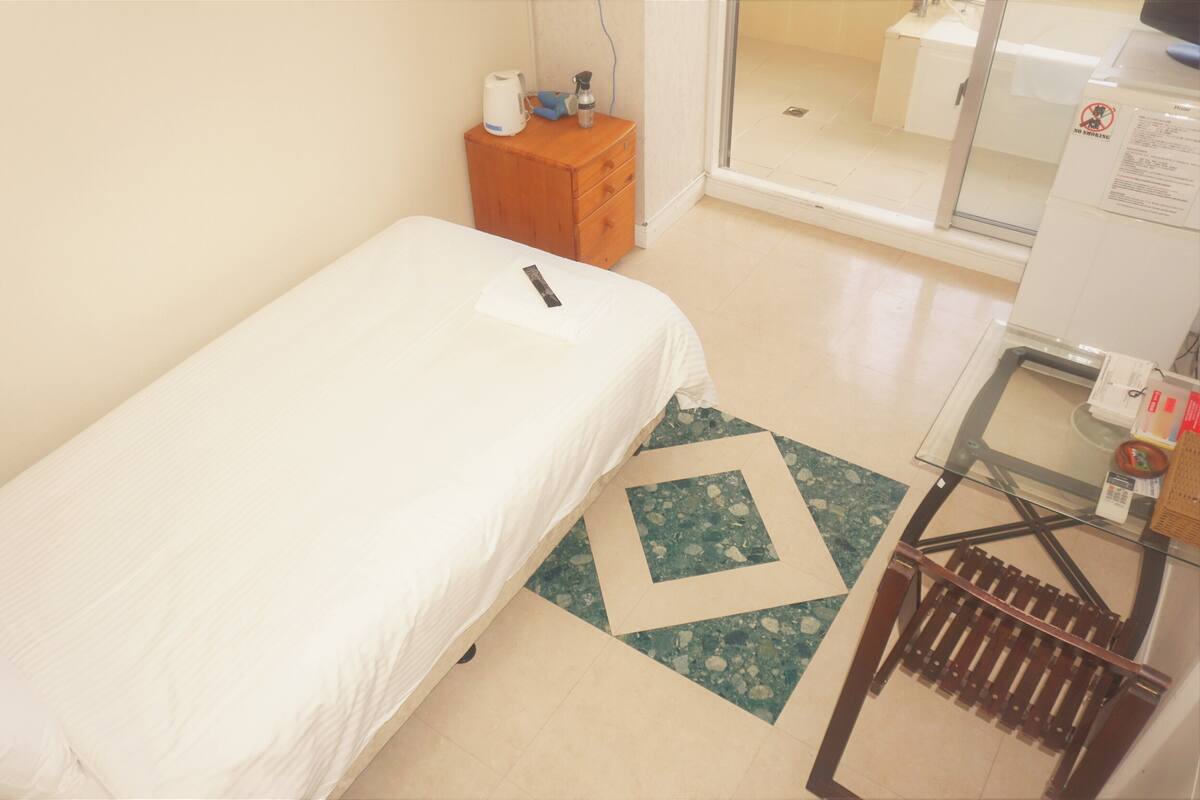 A small, single-bed room with a wooden nightstand, a glass shower enclosure, and a tiled floor.