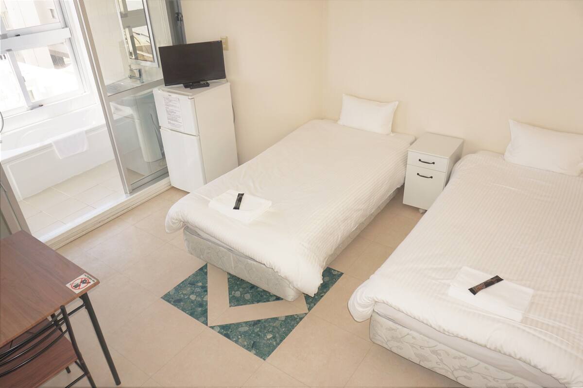 A hotel room with two single beds, a desk, a chair, a television on a cabinet, and a bathroom with a glass door.