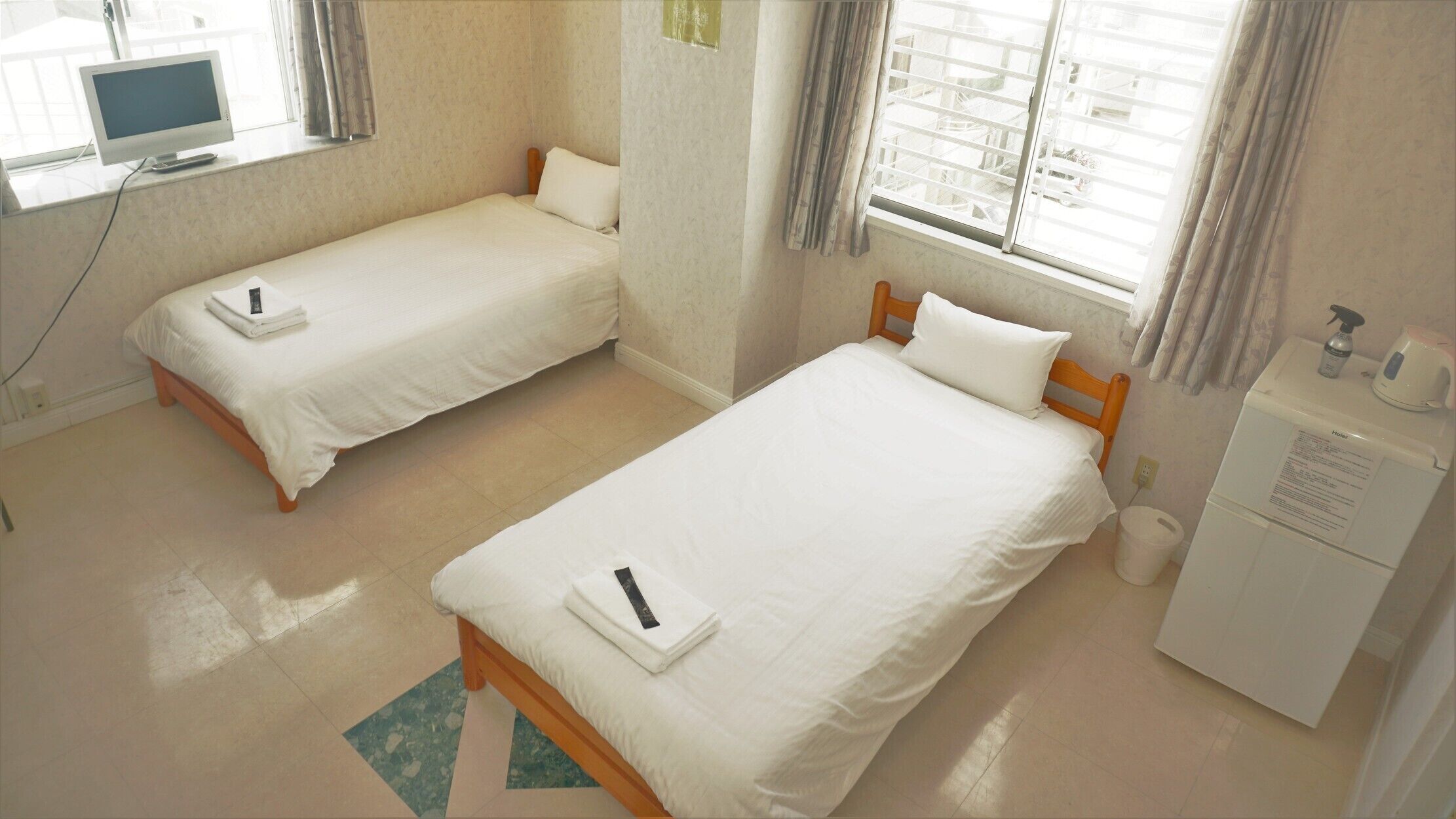 A hotel room with two single beds, a desk with a computer, a heater, and a window with curtains.