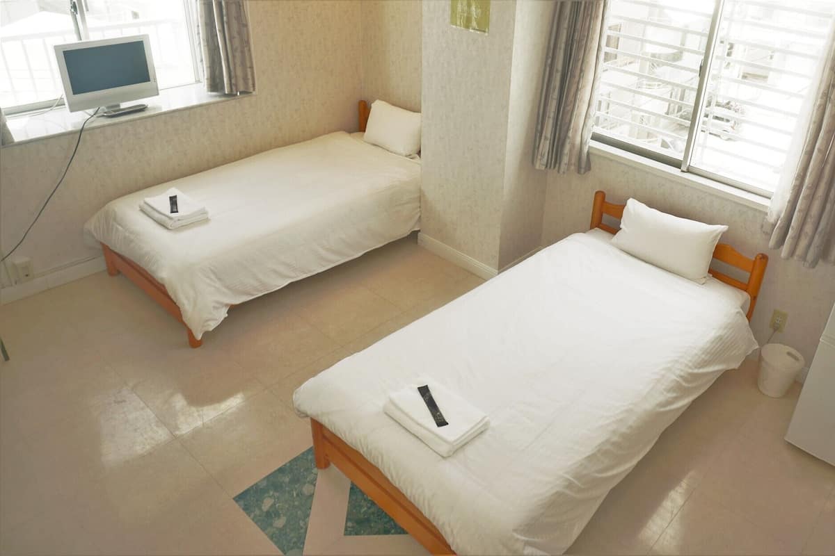 A hotel room with two single beds, a desk with a computer, a heater, and a window with curtains.