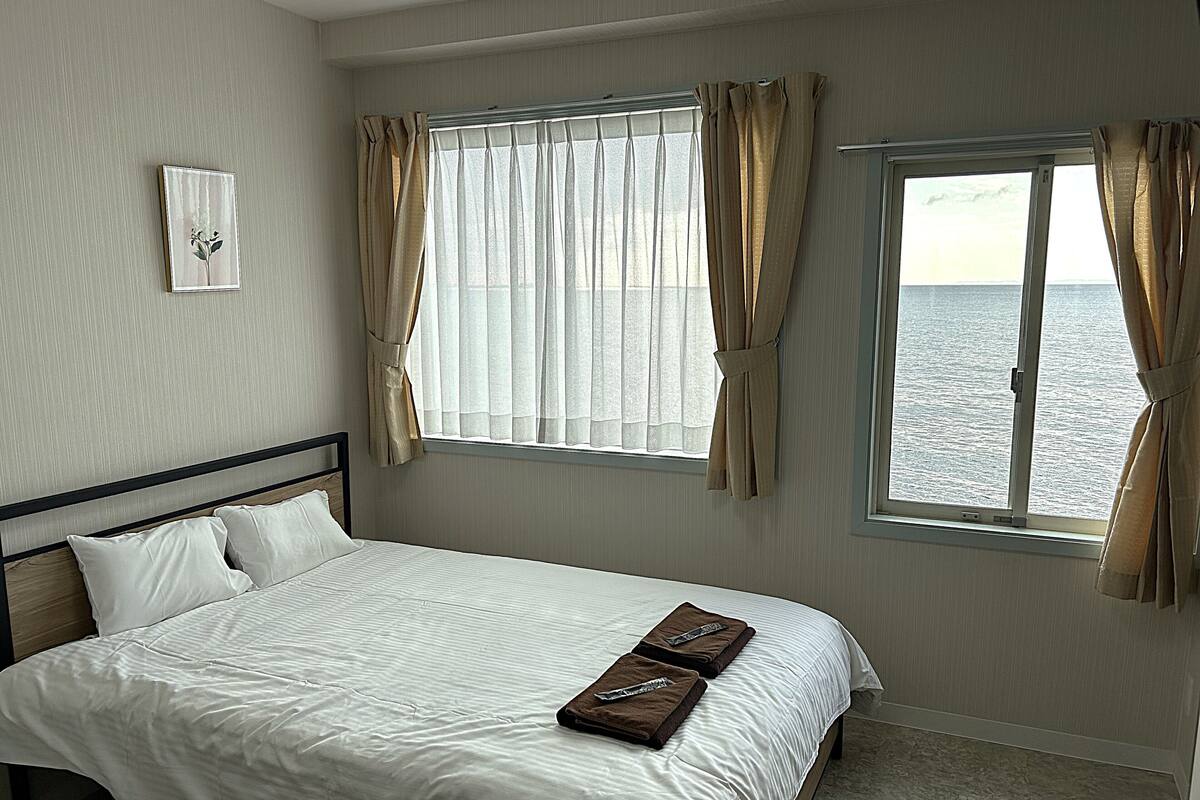 A hotel room with a bed, a window with curtains, a view of the sea, and a framed picture on the wall.