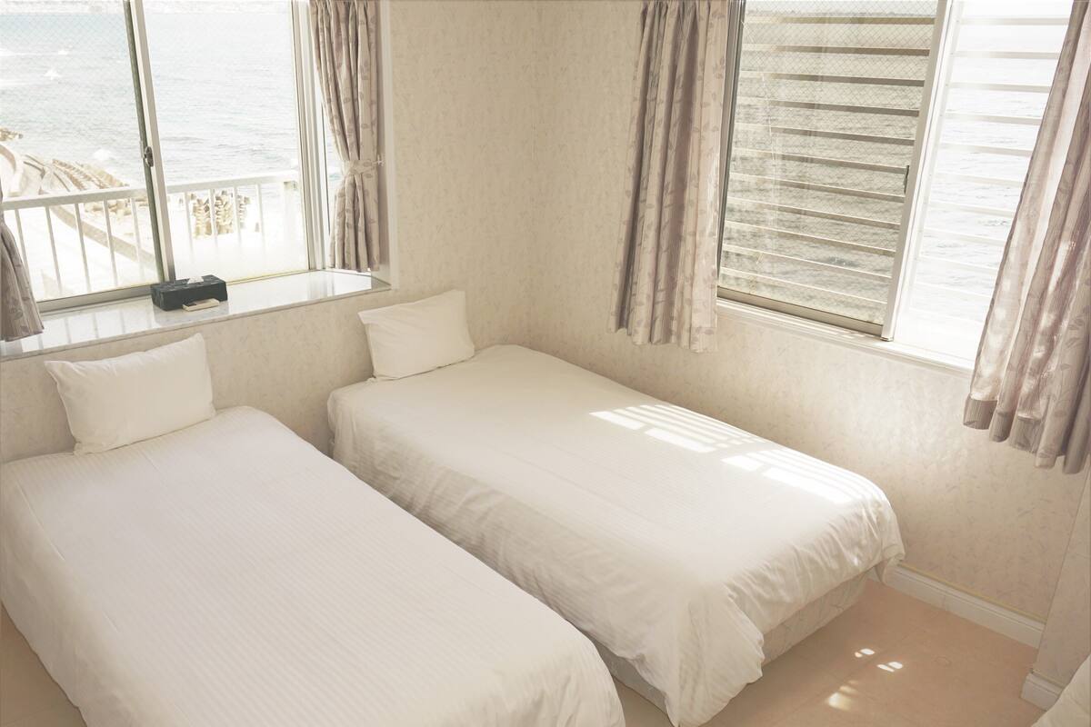 A bedroom with two beds, a window with curtains, and a view of the sea.