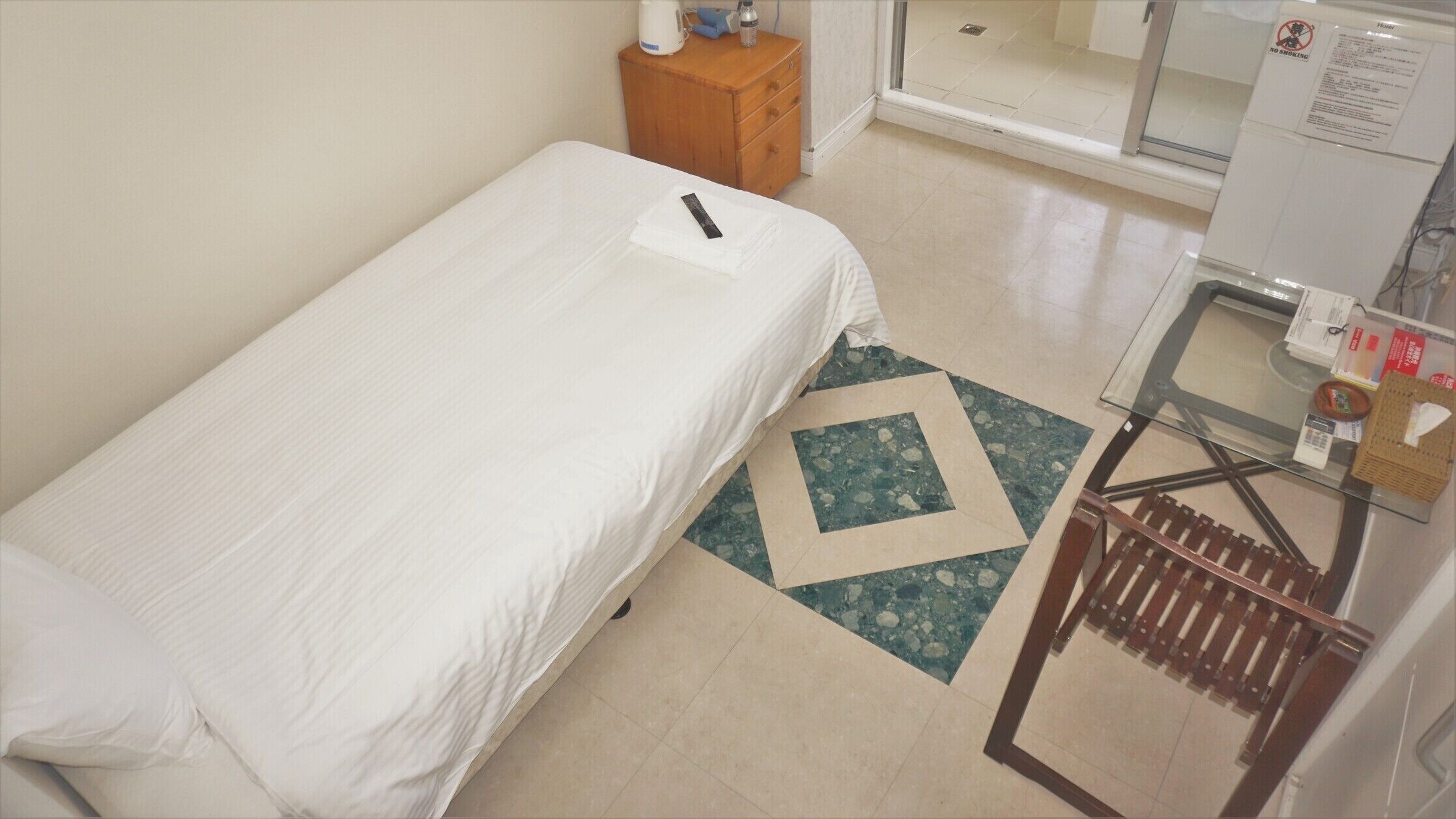 A single bed with a white sheet, a wooden nightstand with a kettle and a bottle, a small table with a basket, and a 'NO SMOKING' sign.