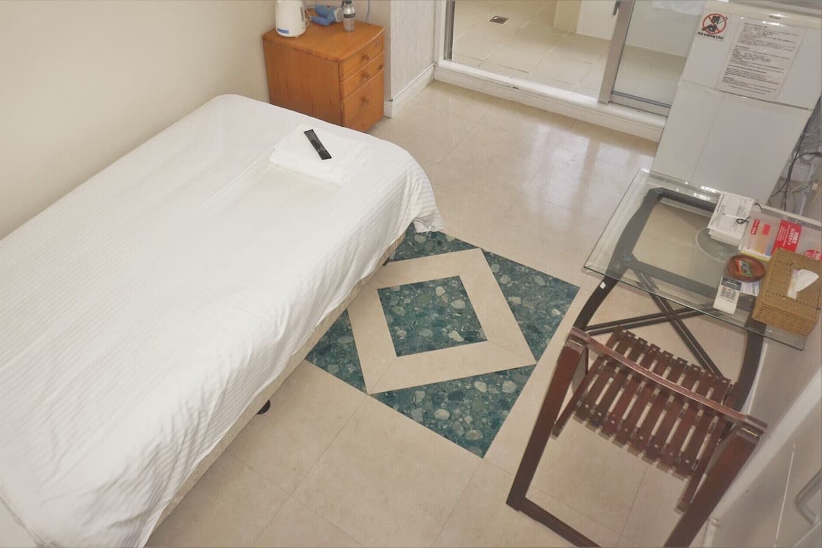 A single bed with a white sheet, a wooden nightstand with a kettle and a bottle, a small table with a basket, and a 'NO SMOKING' sign.