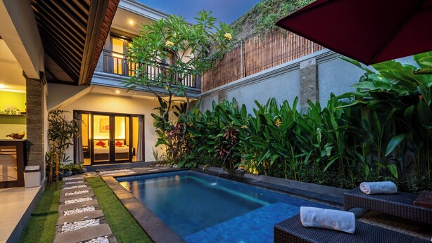 A modern outdoor pool area with a stone pathway, lounge chairs, and lush greenery. - De'bharata Bali Villas Seminyak (Seminyak)