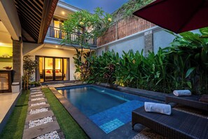 A modern outdoor pool area with a stone pathway, lounge chairs, and lush greenery. - De'bharata Bali Villas Seminyak (Seminyak)