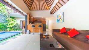 A modern living area with a sofa, a coffee table, and a kitchen in the background. - De'bharata Bali Villas Seminyak (Seminyak)