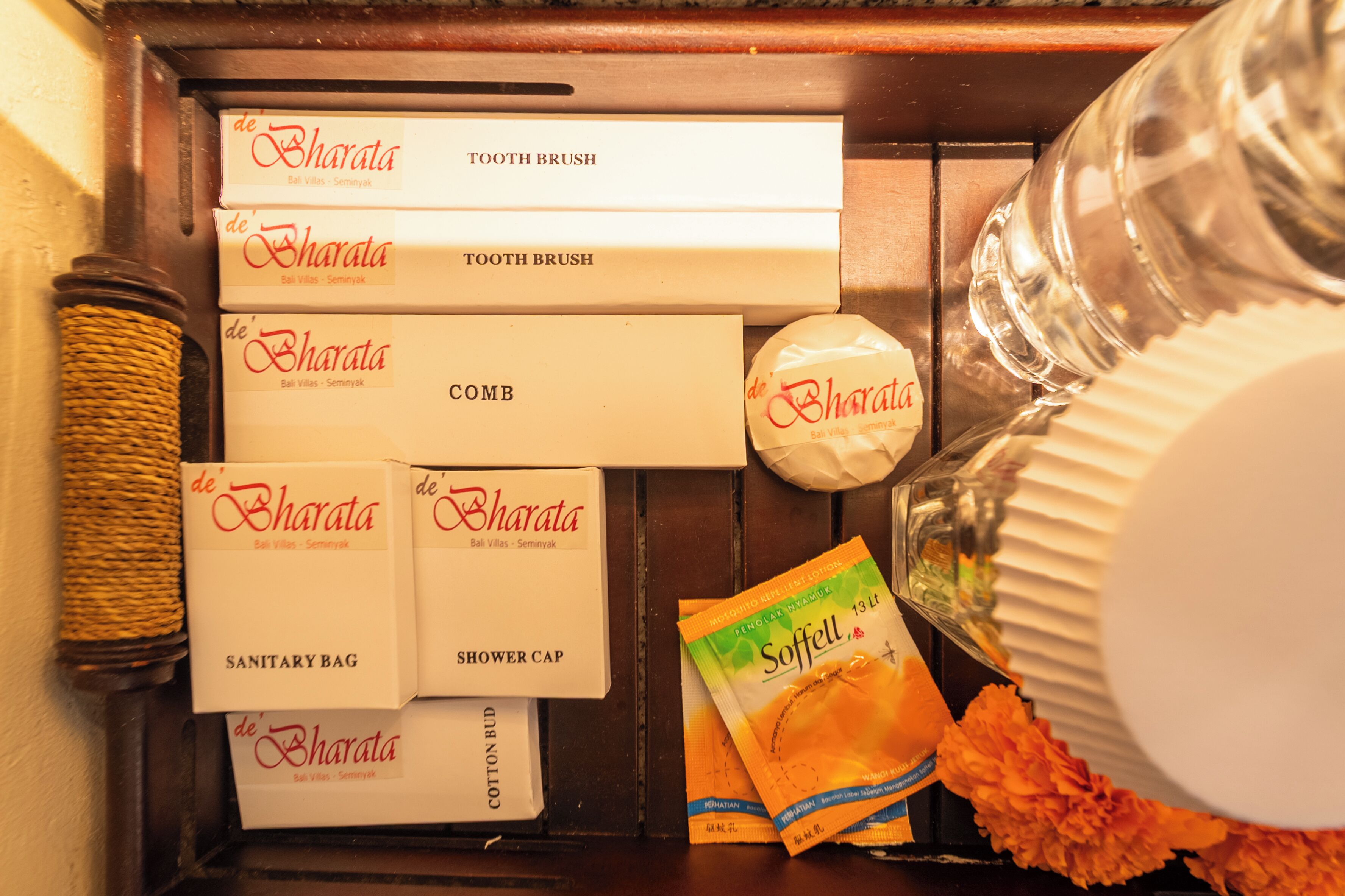 A wooden shelf with various personal care items including toothbrushes, combs, sanitary bags, shower caps, and cotton buds, along with a bottle of Saffell soap and some orange flowers.