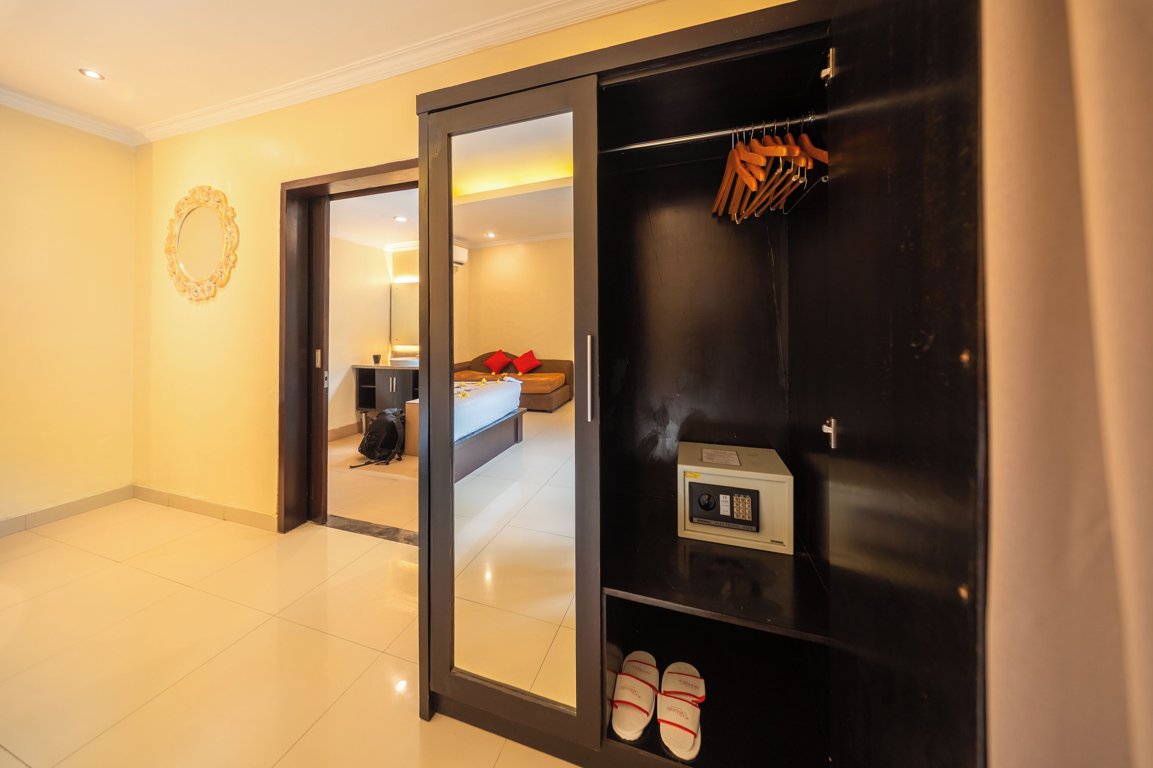 A hotel room with a large mirror, a wardrobe with a shoe rack, and a bed with a red pillow.