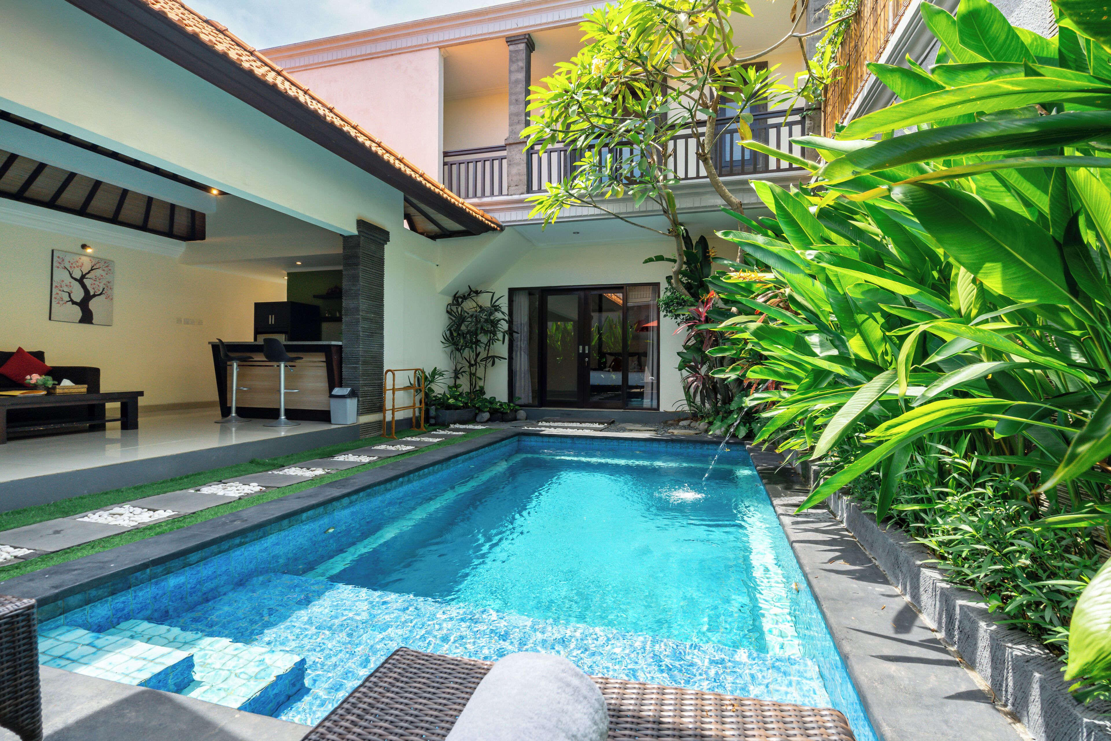 A swimming pool with a tiled deck, surrounded by lush greenery and a two-story building with balconies.
