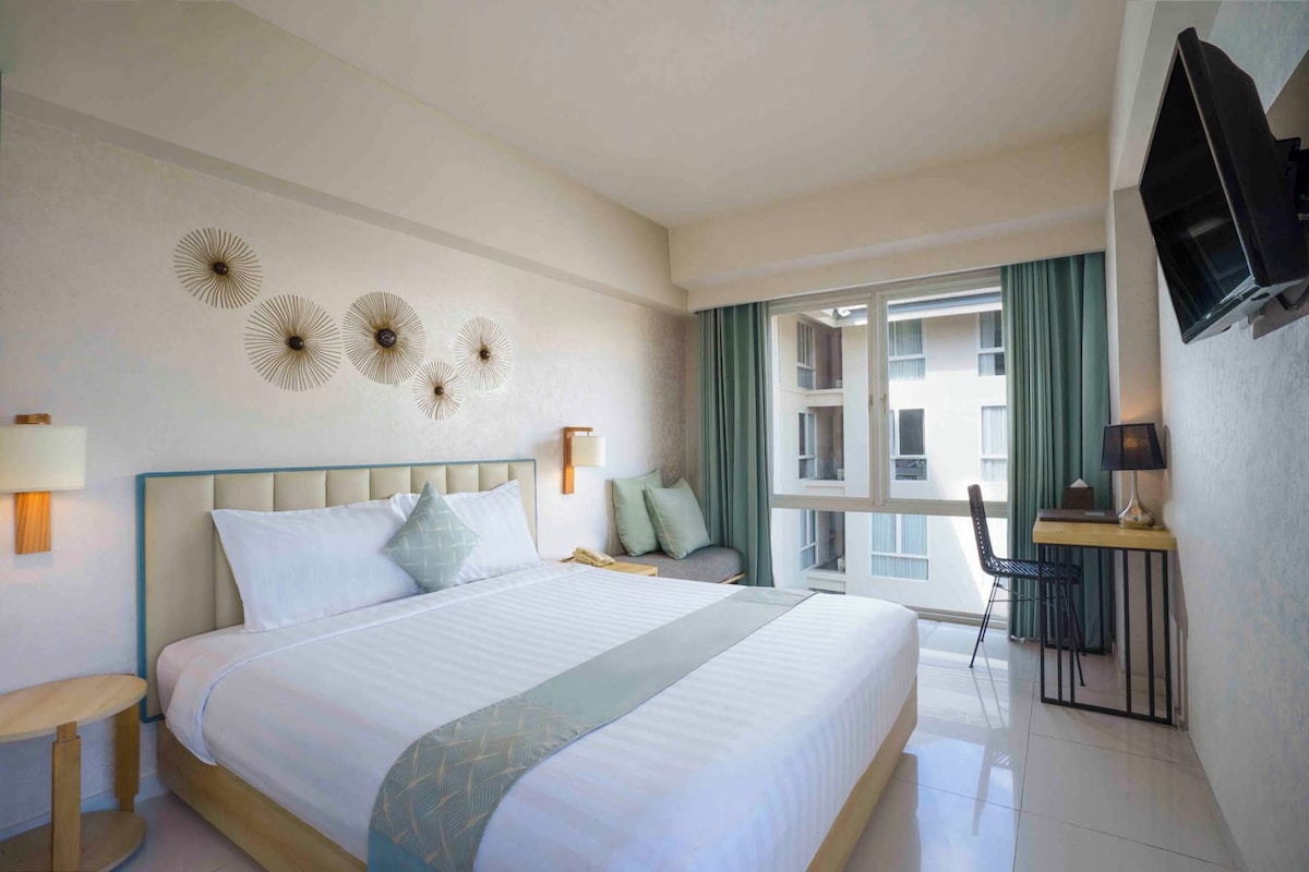 Quarto de hotel com uma cama grande, mesas de cabeceira, uma escrivaninha com cadeira, televisão na parede e uma janela com cortinas.