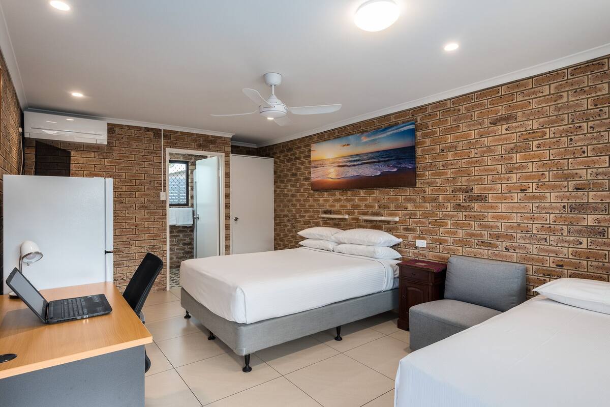 A hotel room with a brick wall, a bed, a desk with a laptop, a chair, and a painting of a sunset.