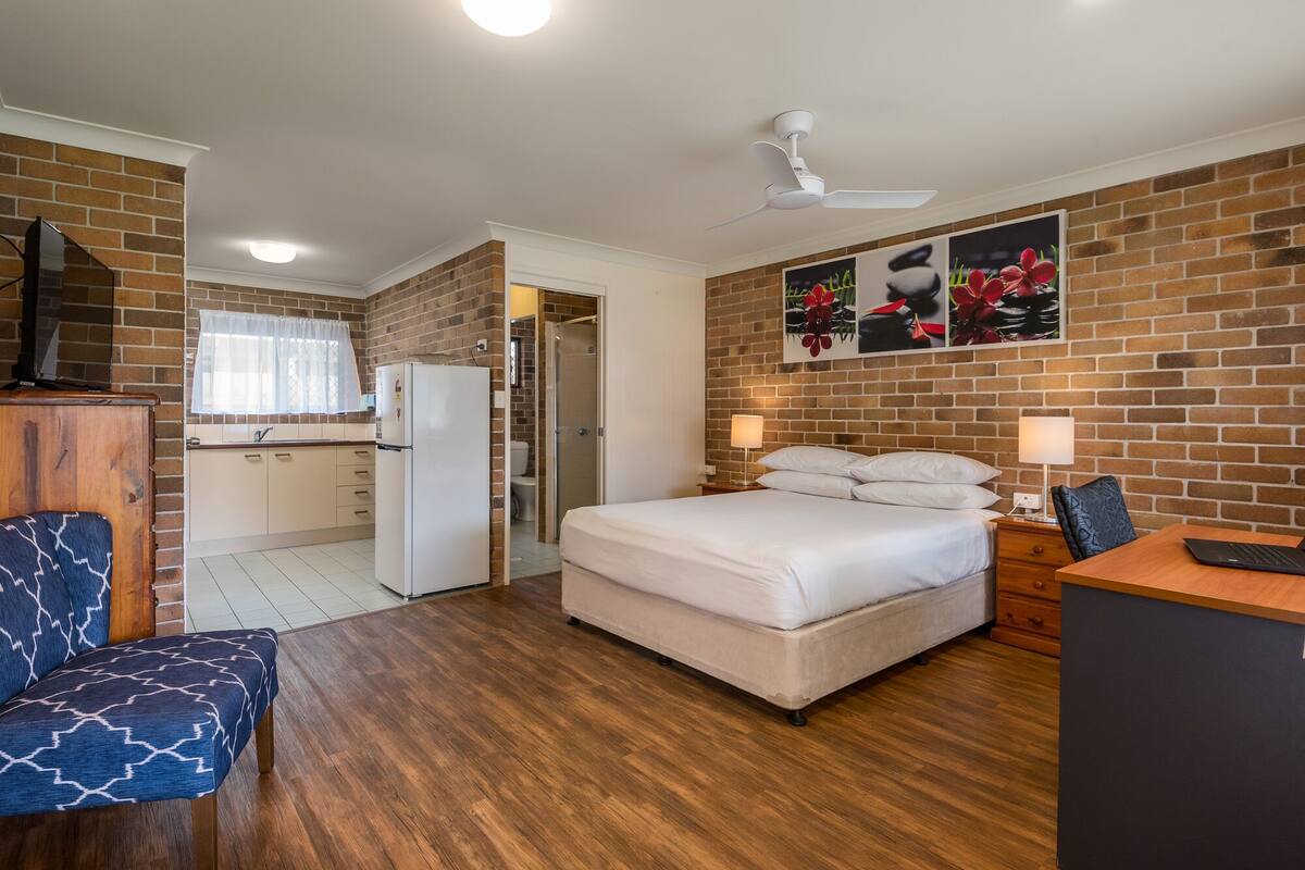 A compact hotel room with a bed, desk, chair, and kitchenette.
