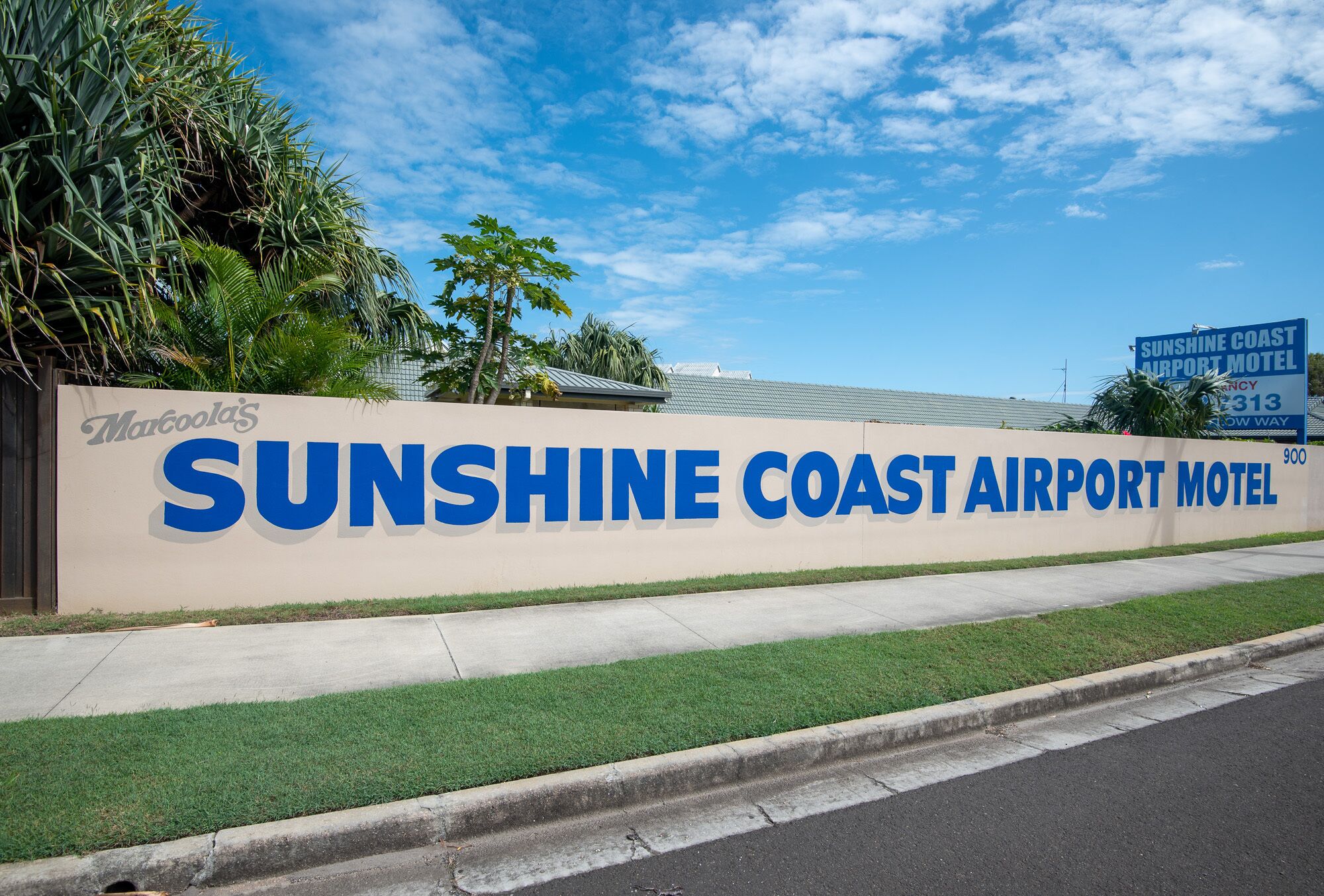 A wall with the Sunshine Coast Airport Motel sign, a concrete sidewalk, and some greenery.