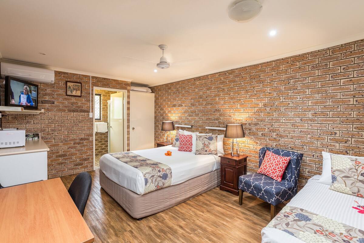 A room with a brick wall, a bed with floral bedding, a desk, and a TV on the wall.