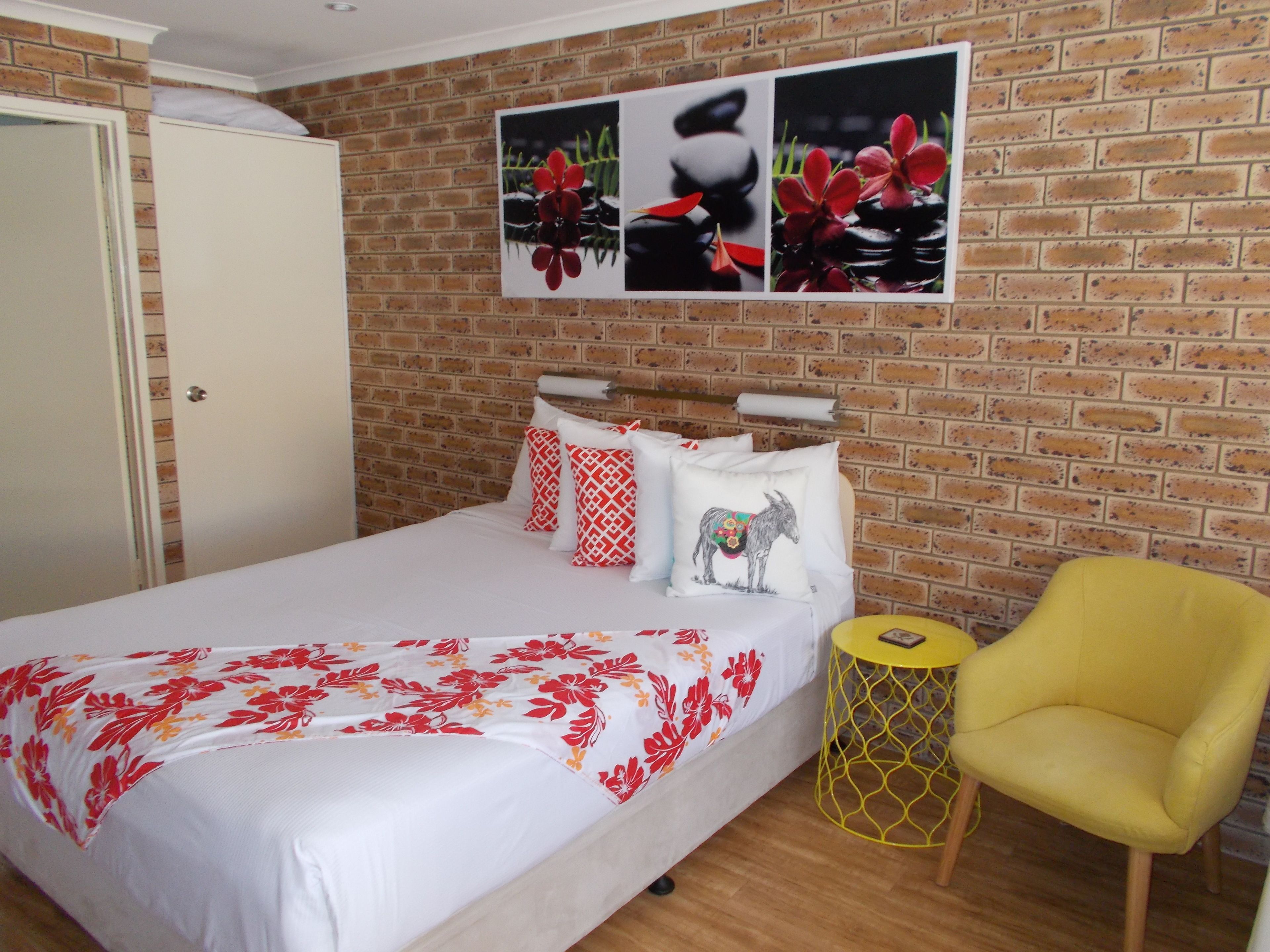 Camera d'albergo con parete in mattoni, un letto con biancheria bianca e motivi floreali rossi, una sedia gialla e un'opera d'arte appesa sopra il letto.