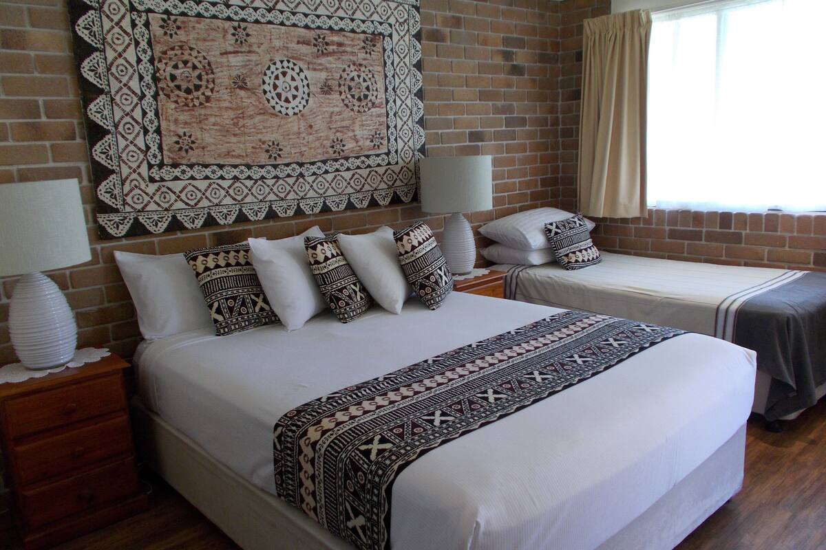 A hotel room with a large bed, two bedside tables, a nightstand, a patterned wall hanging, and a window with curtains.