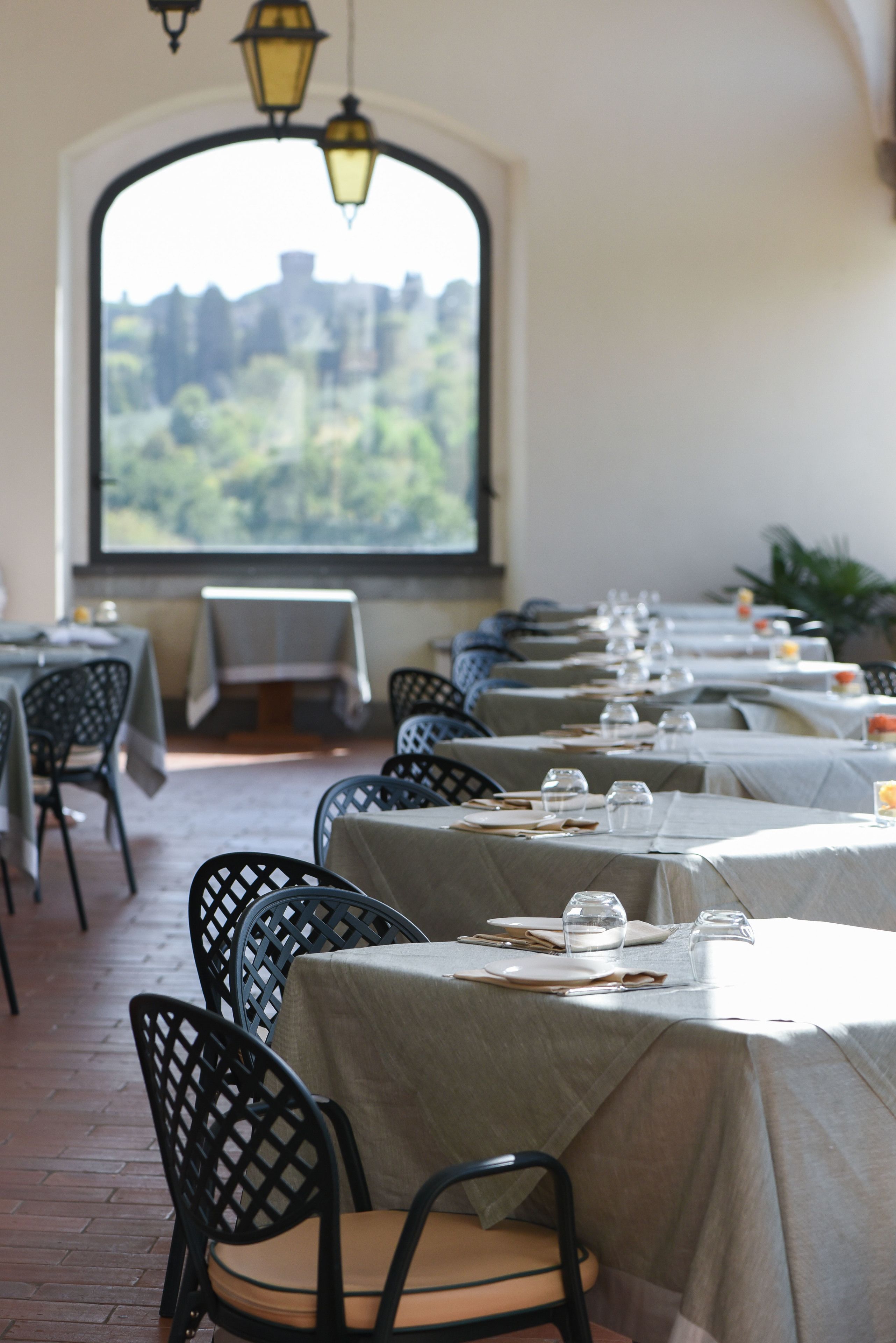 Ein Restaurant mit gedeckten Tischen, Blick auf Grün hinter einem großen Fenster und hängenden Laternen.
